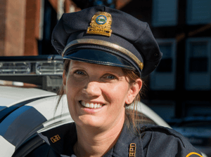 A female police officer in full uniform poses in front of her car and smiles at the camera