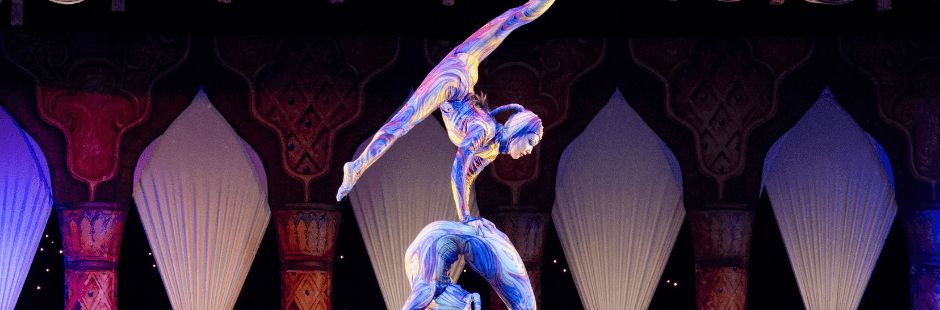 One acrobat balances on top of another, who is bent over in a bridge shape.