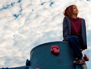 A young boy with red hair sits on a play structure with a sky of puffy white clouds behind him
