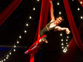 A silk artist hangs on red silk against a black backdrop, with bistro lights