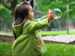 A little girl extends her arms out to pop a soap bubble